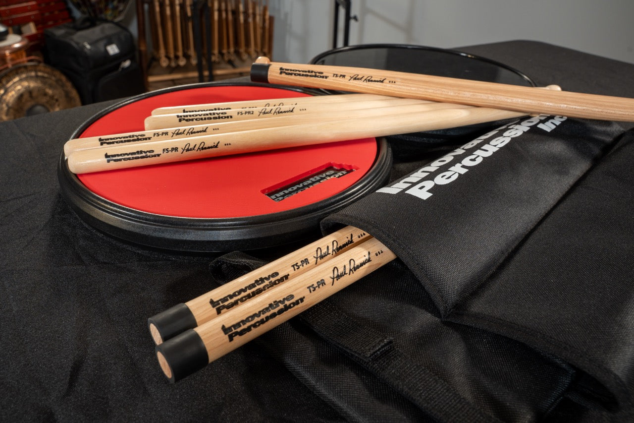 Several pairs of Paul Rennick signature marching drumsticks placed on Innovative Percussion practice pads and a marching stick quiver.